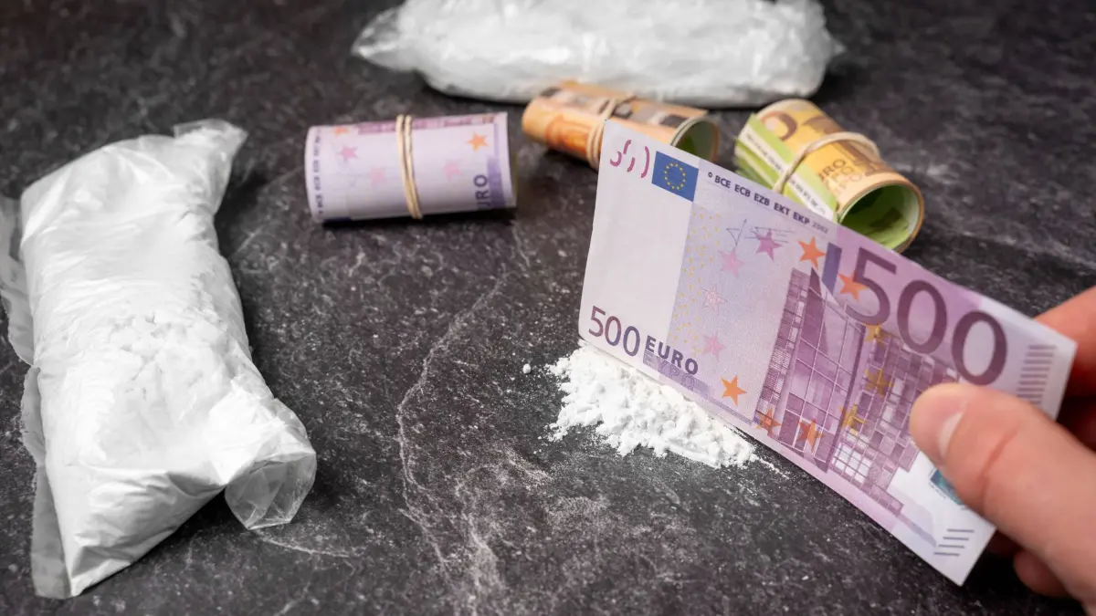 Bavaria, Germany - March 20, 2026: Staged scene - A person is preparing cocaine using a banknote, while additional rolls of cash and small packages drug packets/bags lie in the background. The image symbolizes drug trafficking, drug use, and the connection between illegal business, addiction, and profit. *** Gestellte Szene - Eine Person bereitet Kokain mit einem Geldschein vor, während weitere Geldrollen und Päckchen Drogenpäckchen / Beutel im Hintergrund liegen. Das Bild symbolisiert Drogenhandel, Konsum und den Zusammenhang zwischen illegalem Geschäft, Sucht und Profit.