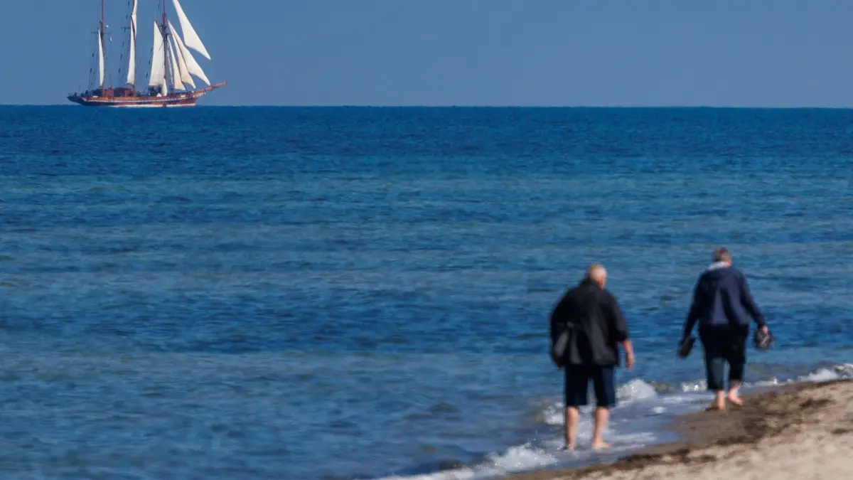 Vorsaison an der Ostseeküste: 19.05.2026, Mecklenburg-Vorpommern, Timmendorf (Poel): Spaziergänger sind am Strand auf der Ostseeinsel Poel unterwegs, während das Traditionsschiff «Amphitrite» auf der Ostsee seine Segel setzt. Foto: Jens Büttner/dpa +++ dpa-Bildfunk +++