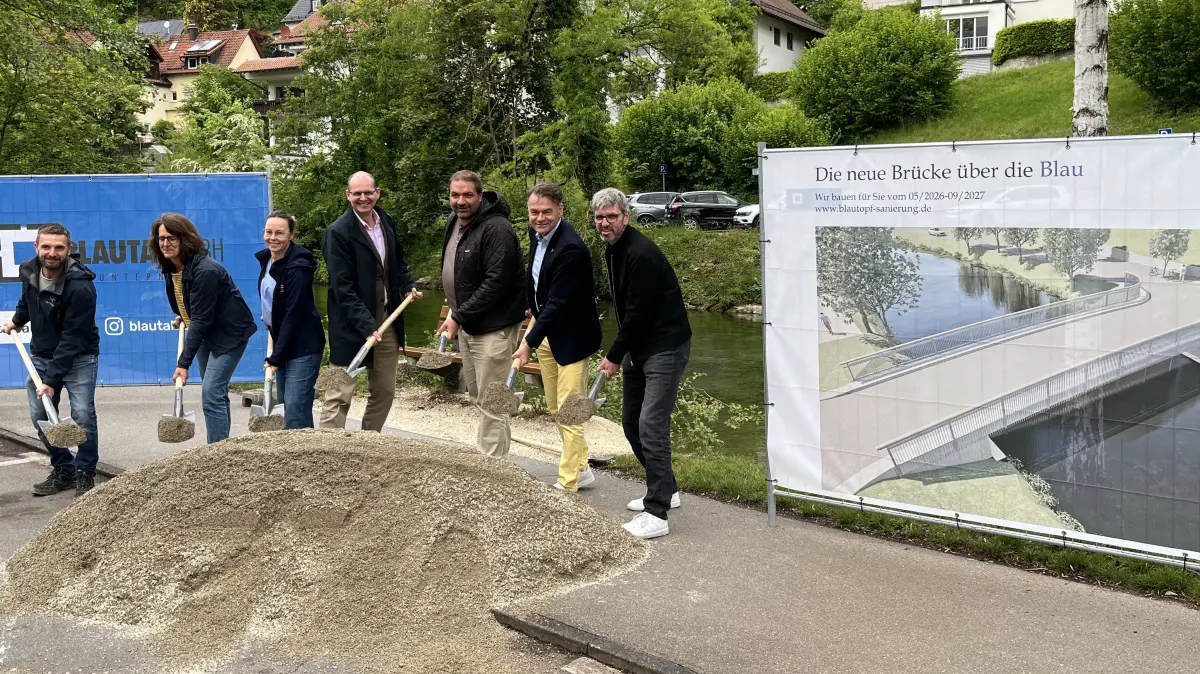 Auftraggeber, Planer und Firmenmitarbeiter zeigen beim offiziellen Spatenstich für den Baustart an der Brücke, wo sich künftig der neue Übergang über die Blau befindet.