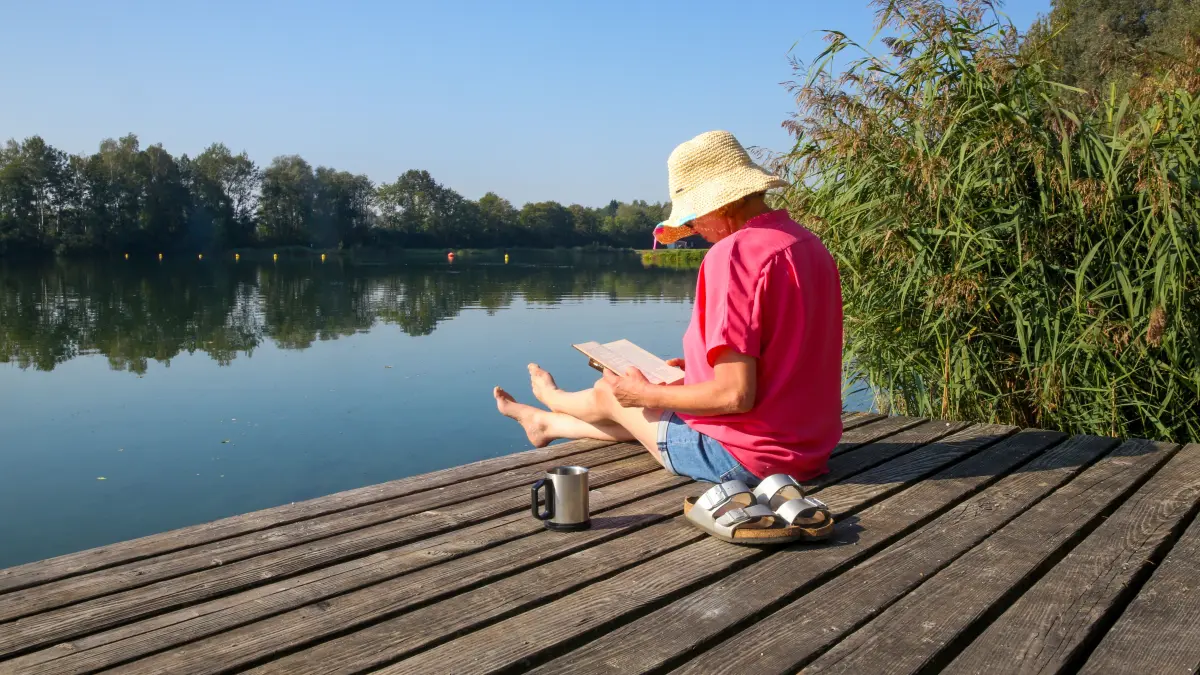Eine Frau sitzt mit einem Buch in der Hand und einer Tasse Kaffe auf einer Badeplattform in der Sonne. +++ dpa-Bildfunk +++ (Symbolbild)