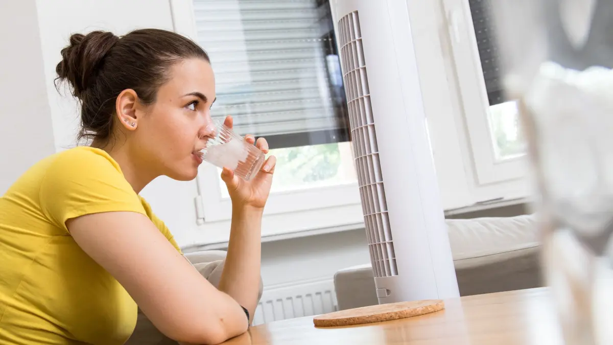 Eine Frau kühlt sich mit Wasser und Ventilator ab: ILLUSTRATION - Viel trinken und bewegte Luft: Damit kann man heiße Tage besser überstehen. (zu dpa: «Heiße Wohnung: Räume clever kühl halten ohne Klimaanlage») Foto: Christin Klose/dpa-tmn - Honorarfrei nur für Bezieher des dpa-Themendienstes +++ dpa-Themendienst +++