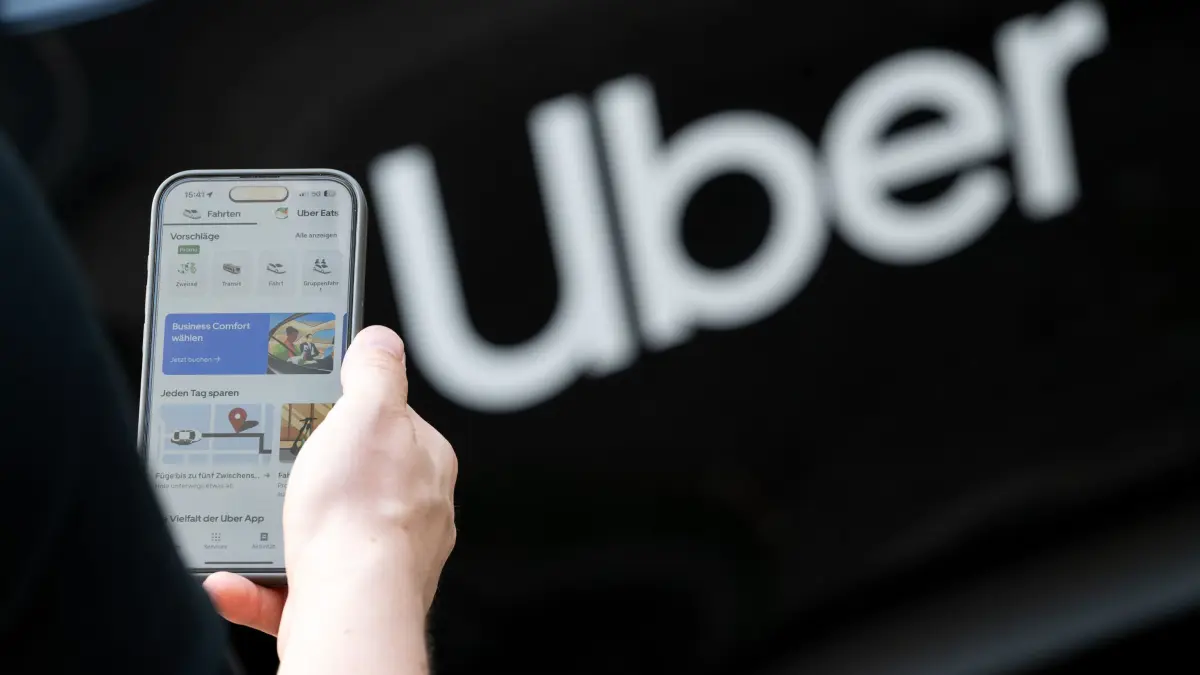 Uber: ILLUSTRATION - 08.08.2024, Berlin: Ein Nutzer steht vor einem Mietwagen mit einem Schriftzug des Mobilitätsdienstleisters und Fahrtenvermittlers Uber und hält sein Smartphone mit der App des Unternehmens in der Hand. (zu dpa: «Uber erlaubt Tonaufnahmen – neue Sicherheitsfunktion startet») Foto: Sebastian Gollnow/dpa +++ dpa-Bildfunk +++
