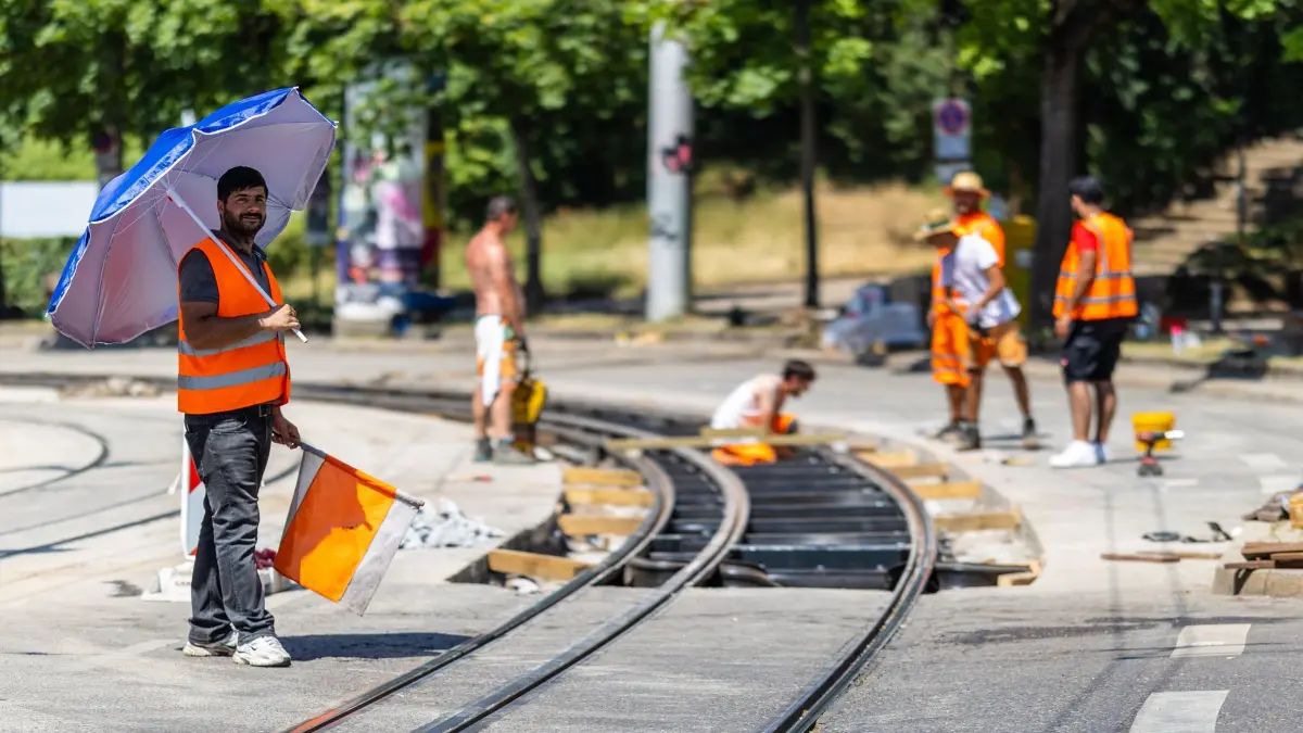 Hitze in Deutschland - Baden-Württemberg: 26.06.2026, Baden-Württemberg, Stuttgart: Ahmed steht bei starker Hitze mit einem Sonnenschirm an einer Baustelle. Er ist dort dafür verantwortlich, dass Autos nicht in das Gleisbett fahren. Die Straße im Stuttgarter Osten wird sowohl von der Stadtbahn, also auch von Autos genutzt - aktuell darf wegen Bauarbeiten allerdings nur die Stadtbahn dort verkehren. Ahmed und seine Kollegen müssen daher in der glühenden Hitze dafür Sorgen, dass keine Autofahrer aus Versehen in das offene Gleisbett fahren. Foto: Christoph Schmidt/dpa +++ dpa-Bildfunk +++