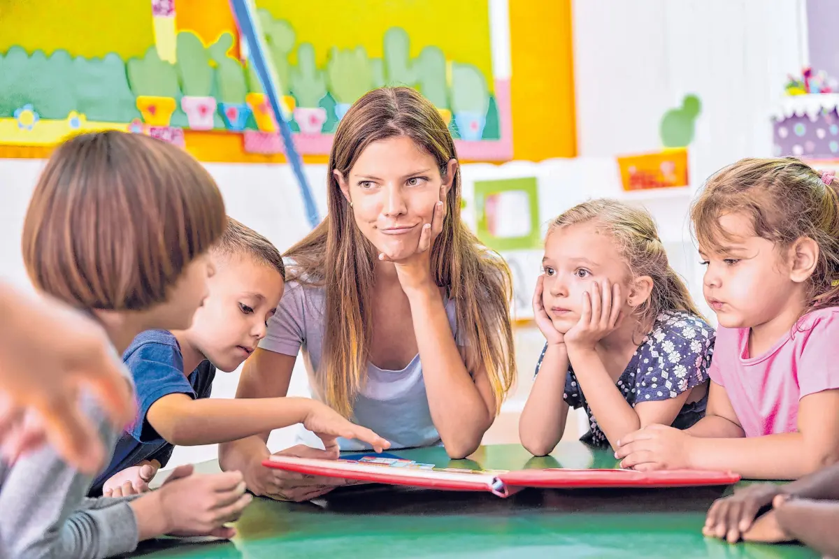 Tagesmutter oder Kindergärtnerin und Kinder lesen und spielen zusammen im Kindergarten⇥Kneschke
