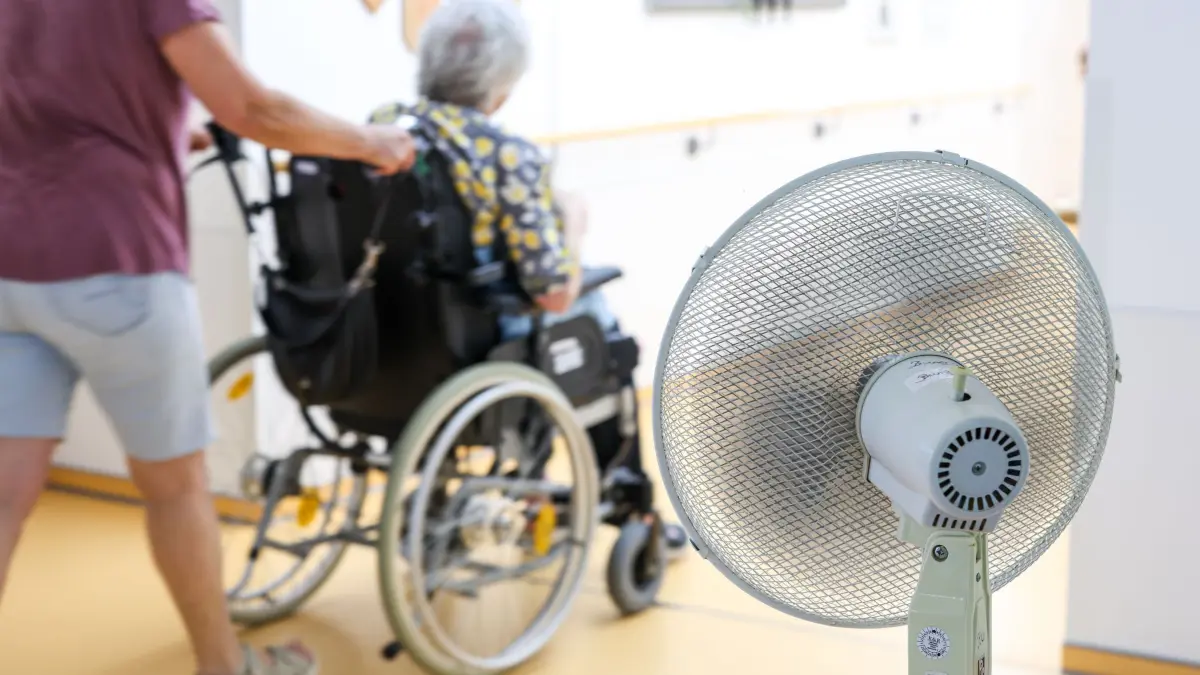 Ältere Menschen leiden unter hohen Temperaturen: ARCHIV - 25.06.2026, Nordrhein-Westfalen, Grefrath: Eine Mitarbeiterin eines Seniorenheims schiebt eine Bewohnerin über den Flur, während ein Ventilator einen kühlen Luftzug erzeugt. (zu dpa: «RKI-Schätzung: Tausende Tote durch Hitzewelle Ende Juni») Foto: Christoph Reichwein/dpa +++ dpa-Bildfunk +++