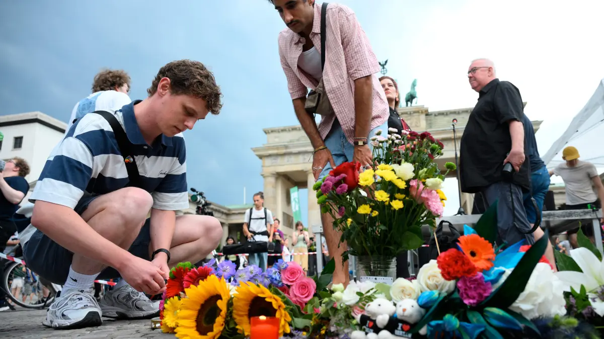 GERMANY-LGBTQ+-PARADE-CSD-POLICE: Mourners place bouquets of flowers on July 26, 2026 in front of landmark Brandenburg Gate during to a so-called "community mourning" one day after the car ramming attack on people on the outskirts of Berlin's Christopher Street Day (CSD) parade on late July 25, 2026, killing one person and injuring 16 in the Tiergarten park. German police searched on July 26, 2026 for a suspect believed linked to Islamist circles after a car ploughed into a crowd near Berlin's Gay Pride celebrations, killing one person and wounding 16 others. The incident shattered what had been a day of celebration for hundreds of thousands attending Christopher Street Day, one of Europe's largest Pride events, and left revellers fleeing a central Berlin park as sirens echoed through the night. (Photo by RALF HIRSCHBERGER / AFP)