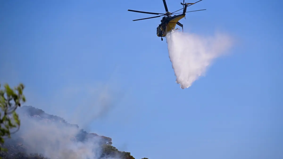 Waldbrände in Griechenland: ARCHIV - 09.08.2025, Griechenland, Charvalo: Ein Löschhubschrauber wirft nach einem Waldbrand im Dorf südöstlich von Athen Wasser auf ein wieder aufflammendes Feuer. (zu dpa: «Waldbrand auf Kreta - zwei Feuerwehrleute kommen ums Leben») Foto: Michael Varaklas/AP/dpa +++ dpa-Bildfunk +++