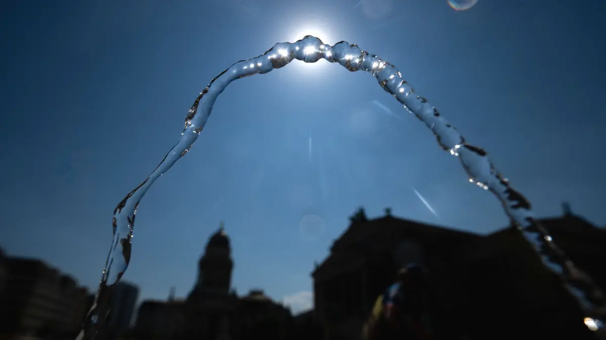 Wetter in Berlin: 30.07.2026, Berlin: Wasser fließt an einem öffentlichen Trinkbrunnen auf dem Gendarmenmarkt in einem Bogen vor der Sonne. Foto: Sebastian Christoph Gollnow/dpa +++ dpa-Bildfunk +++
