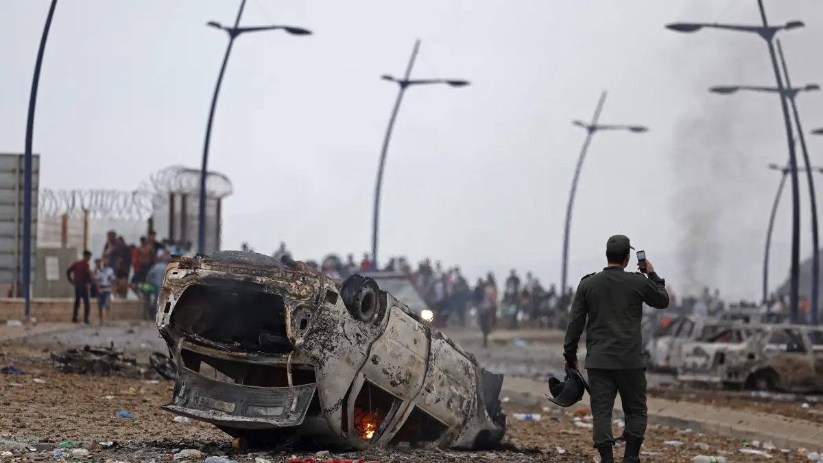 MOROCCO-SPAIN-MIGRATION: A member of Moroccan security forces uses his mobile phone next to an overturned damaged vehicle at the site of clashes near Fnideq, on the Morocco-Spain border, on July 31, 2026. Thousands of migrants crossed overnight from Morocco into Spain's north Africa's Ceuta enclave, a police source said on July 31, adding that it was impossible to estimate how many people in total. A total of 18 people died while attempting to reach the Spanish enclave, the source said. (Photo by Abdel Majid BZIOUAT / AFP)