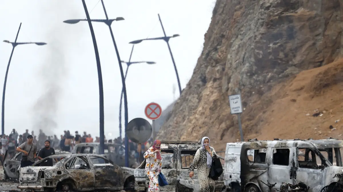 MOROCCO-SPAIN-MIGRATION: People walk past burnt vehicles at the site of clashes near Fnideq, on the Morocco-Spain border, on July 31, 2026. Thousands of migrants crossed overnight from Morocco into Spain's north Africa's Ceuta enclave, a police source said on July 31, adding that it was impossible to estimate how many people in total. A total of 18 people died while attempting to reach the Spanish enclave, the source said. (Photo by Abdel Majid BZIOUAT / AFP)