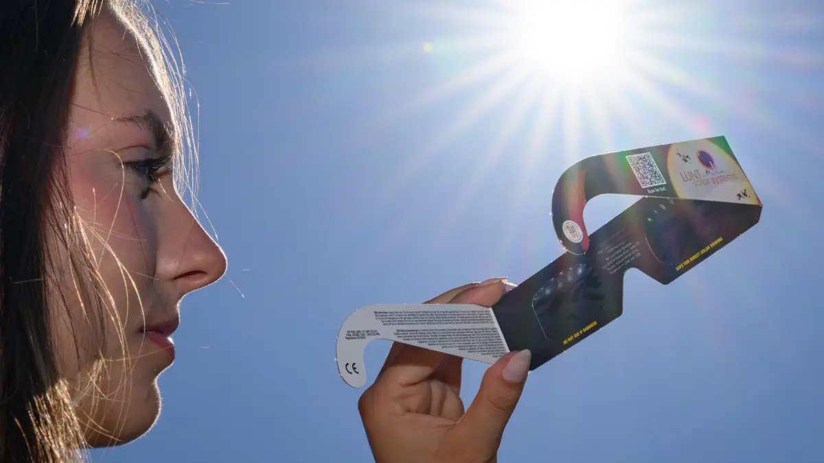 Vorfreude auf die Sonnenfinsternis: 09.08.2026, Brandenburg, Sieversdorf: Eine junge Frau setzt sich eine Sonnenfinsternisbrille auf, um damit die Mittagssonne über Brandenburg zu betrachten. Das Zentralgestirn steht derzeit im Fokus der Astronomie, da am Abend des 12.08.2026 eine bedeutende partielle Sonnenfinsternis stattfindet. Foto: Patrick Pleul/dpa +++ dpa-Bildfunk +++