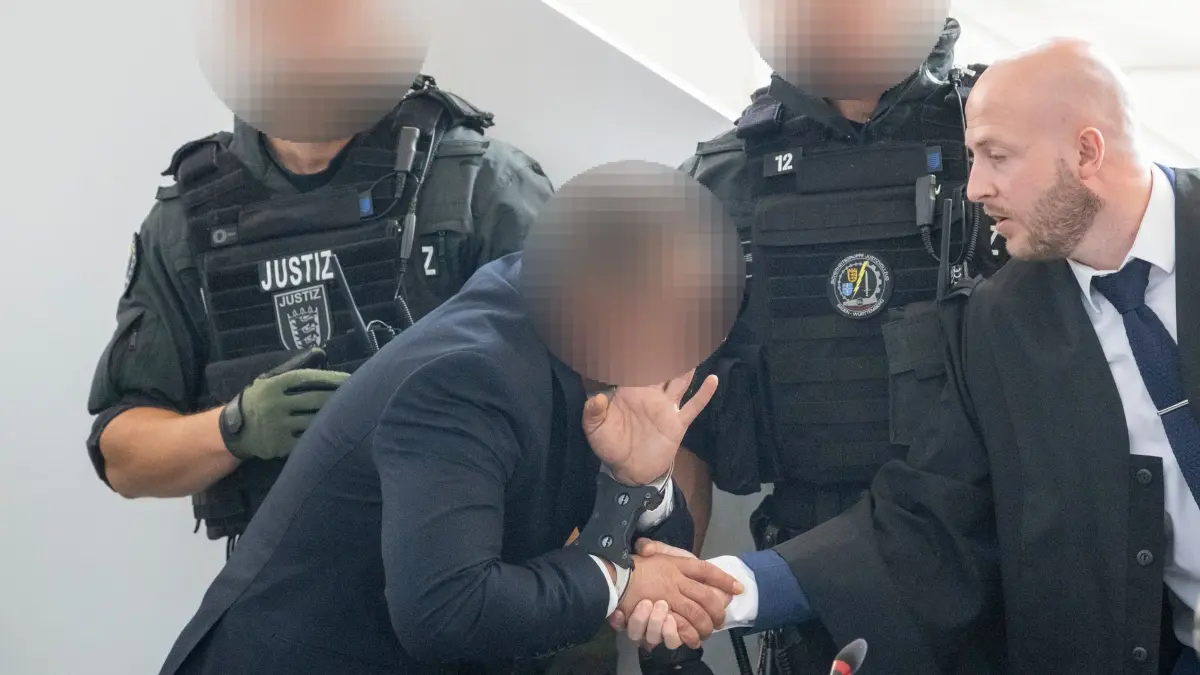 Auftakt Mordprozess Dönerimbiss in Schrozberg: 11.08.2026, Baden-Württemberg, Ellwangen: Justizangehörige bewachen den Angeklagten im Schwurgerichtssaal des Landgerichts. Der Angeklagte gibt seinem Verteidiger Michael Becker die Hand. Ihm wird vorgeworfen, seine Ehefrau mit einer Pistole erschossen zu haben. Foto: Stefan Puchner/dpa +++ dpa-Bildfunk +++