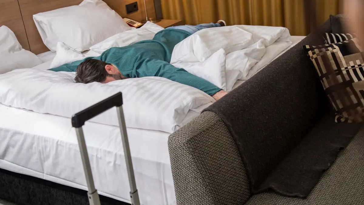 Ein Mann liegt erschöpft auf einem Bett in einem Hotelzimmer: ILLUSTRATION - Kaum angekommen, schon krank: Mit der Entspannung macht nicht selten das Immunsystem schlapp. (zu dpa: «Krank im Urlaub: So holen Sie sich die Urlaubstage zurück») Foto: Christin Klose/dpa-mag - Honorarfrei nur für Bezieher des Dienstes dpa-Magazin +++ dpa-Magazin +++