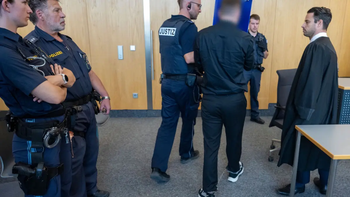 Prozess wegen Mordversuch gegen drei junge Männer: 28.08.2026, Baden-Württemberg, Ulm: Ein Justizwachtmeister führt im Landgericht einen gefesselten Mann zur Anklagebank, wo dessen Anwalt Julian Ackermann (r) auf ihn wartet. Die Staatsanwaltschaft wirft drei jungen Männern vor, gemeinsam einen Mordversuch in einem Imbiss begangen zu haben. Foto: Stefan Puchner/dpa +++ dpa-Bildfunk +++