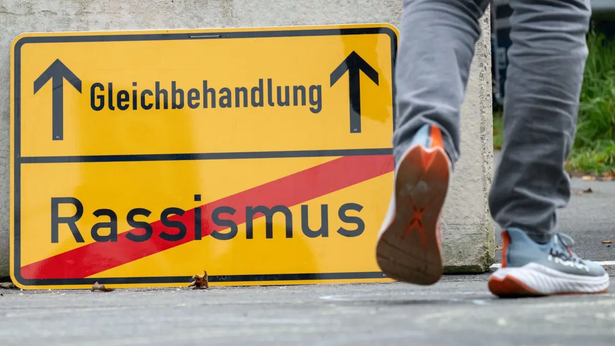 Tag der Deutschen Einheit - Mödlareuth: 03.10.2026, Bayern, Töpen: Ein Besucher läuft auf dem Gelände des Deutsch-Deutschen Museums in Mödlareuth an einem Schild vorbei, auf dem das Wort "Rassismus" (unten) rot durchgestrichen und das Wort "Gleichbehandlung" mit Pfeilen nach oben versehen ist. Anlässlich des Tags der Deutschen Einheit finden in dem ehemals geteilten Ort, der auch als "Little Berlin" bekannt ist, mehrere Veranstaltungen statt. Foto: Pia Bayer/dpa +++ dpa-Bildfunk +++